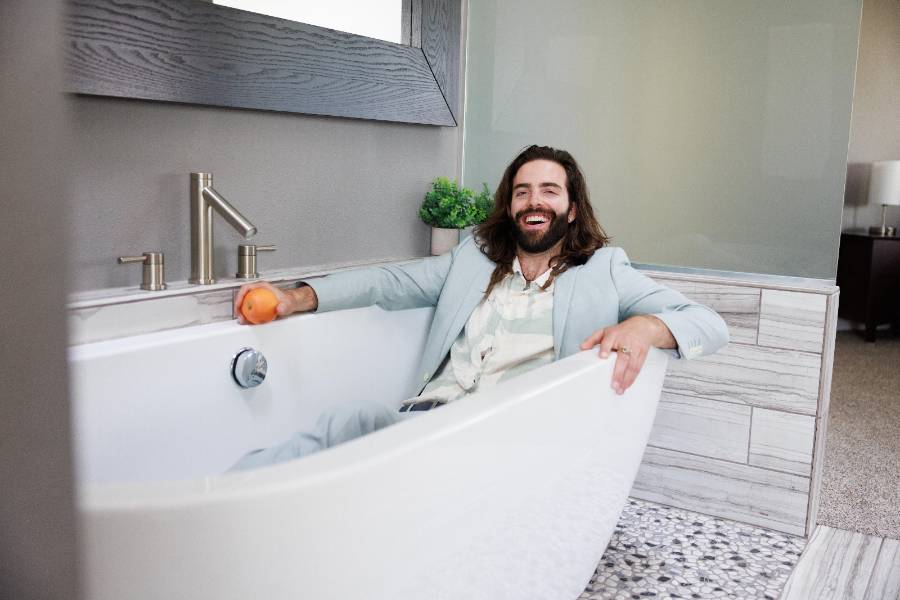 Scott West laughing while sitting fully dressed in an empty bathtub, holding an orange